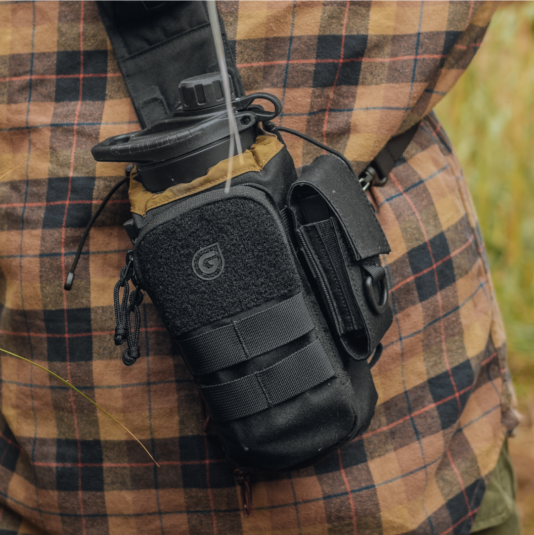 Hiker in plaid shirt carrying Mission Bottle Carrier with G-MOD Multi-Tool Pouch in Covert Black colorway. GeoPress water filter and purifier bottle visible in main compartment.