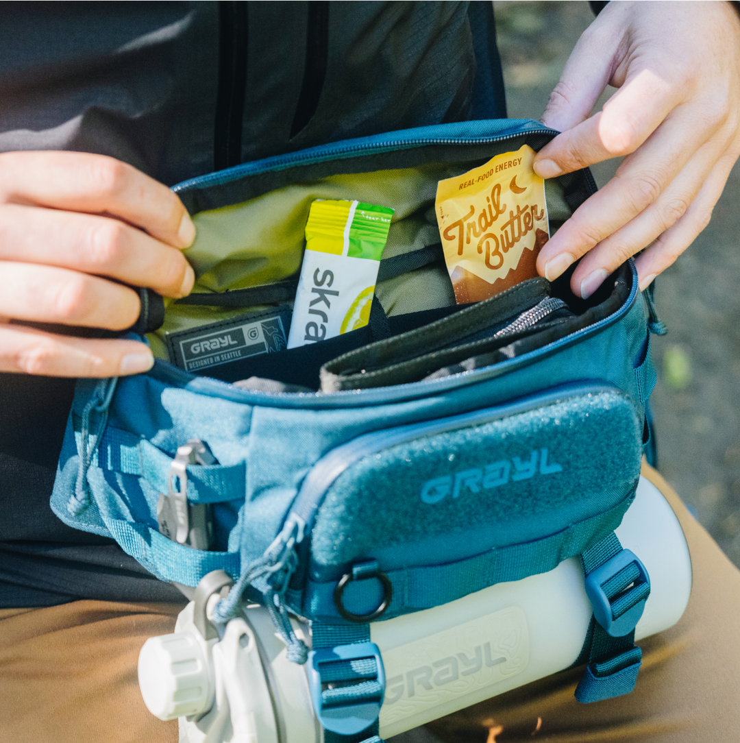 Interior photo of main compartment of 2L Grayl Transport Hip Pack. Trail Butter single serving visible alongside Skratch Labs Electrolyte Drink Mix. GeoPress Alpine White bottle suspended in BottleLock Tie Down System.