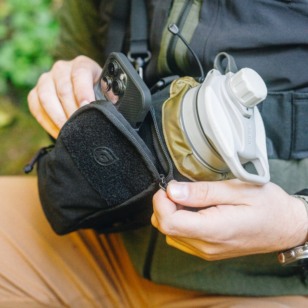 Alpine White GeoPress on its side in main compartment of Transport Bottle Carrier secured by dual-cinch closure. Hiker placing full-size cell phone in pocket.