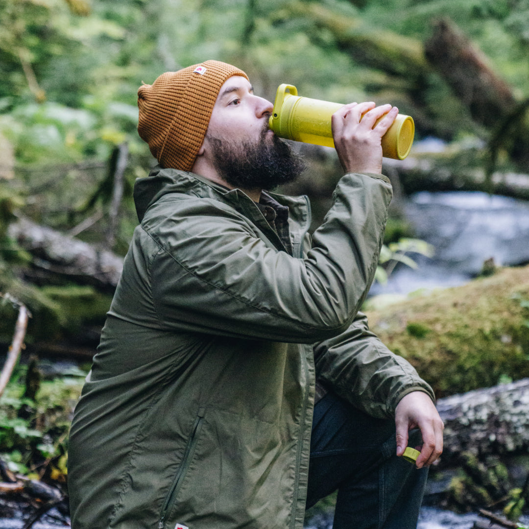 bearded man drinking from his Grayl while leaning on fallen logs with a wooded stream in the background